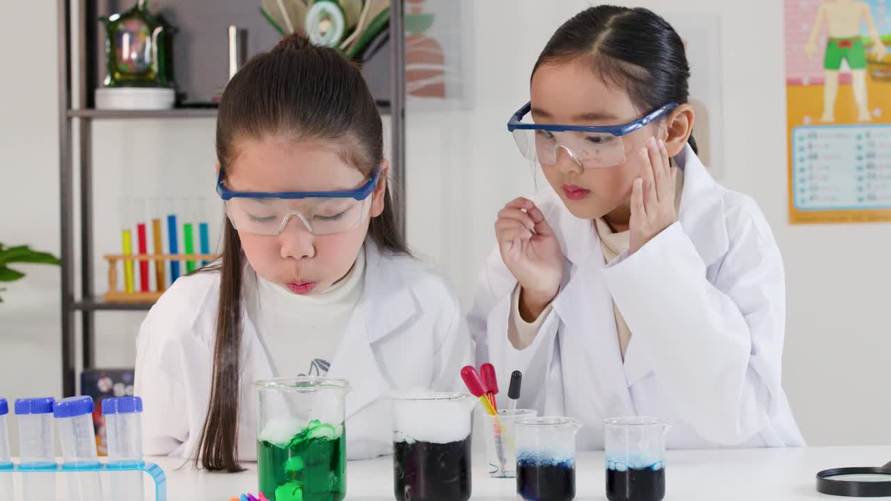 明るい教室で、保護メガネを着用した若いアジア人の女の子2人が化学反応を観察している。