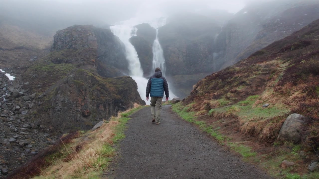 Hiker walking on a path towards the powerful Rjukandafoss waterfall on a foggy day in the Icelandic highlands landscape