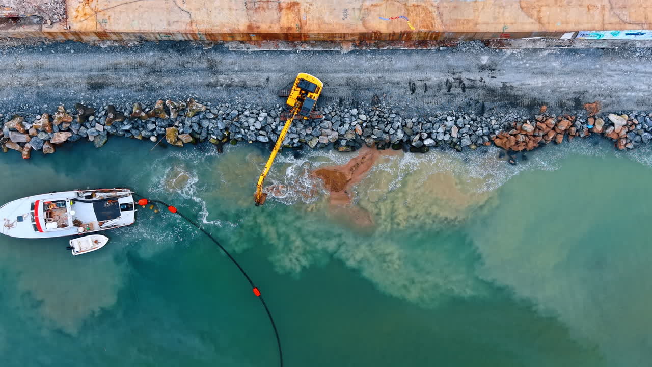 Vista aerea dall'alto di un escavatore al lavoro su una costa rocciosa. Escavatore giallo su un molo in pietra vicino a una barca con sedimenti in acqua turchese.