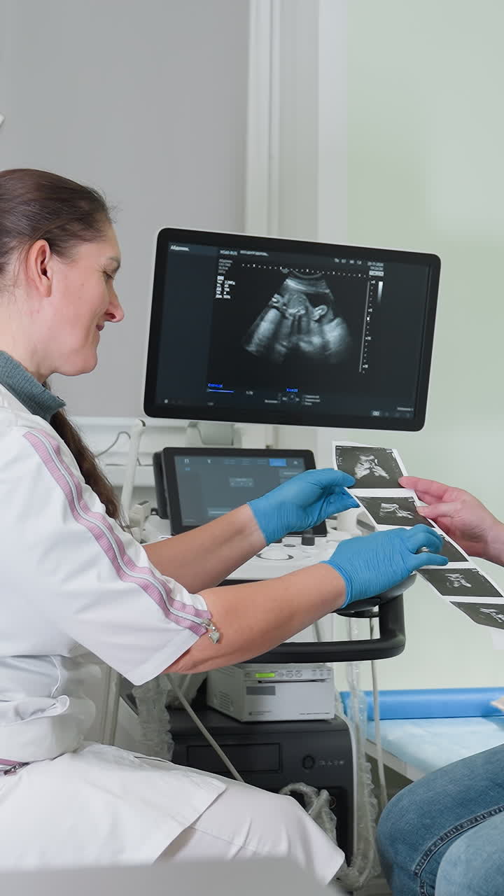 Ecografista caucasico che esegue un'ecografia prenatale, mani con guanti che azionano il trasduttore e la console, monitor che mostra immagini fetali, paziente che riceve foto stampate, ambiente clinico soft