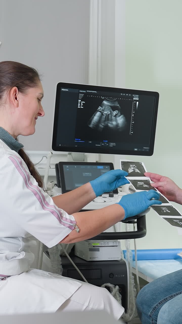 Ecografista caucasico che esegue un'ecografia prenatale, mani con guanti che azionano il trasduttore e la console, monitor che mostra immagini fetali, paziente che riceve foto stampate, ambiente clinico soft