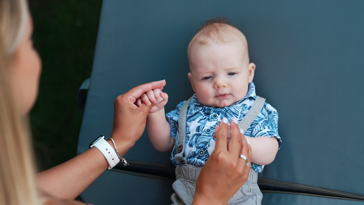 Mom is holding tiny infant's hand moving them to the sides. Mother is playing with her baby son outdoors.