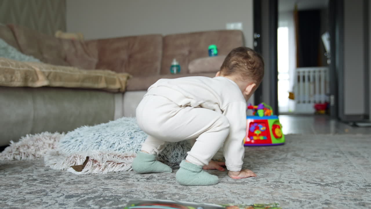 Rear view of a baby in white suit sitting on the floor and eating a cookie. Kid drops the food on the floor, stands up to get it back and eat.