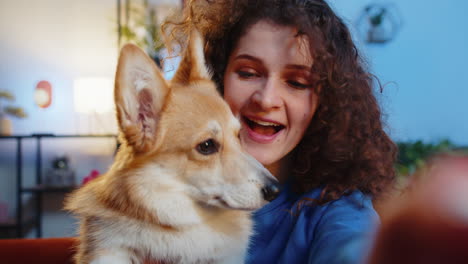 pov of young smiling woman kissing cute corgi dog and taking selfie via smartphone at home room