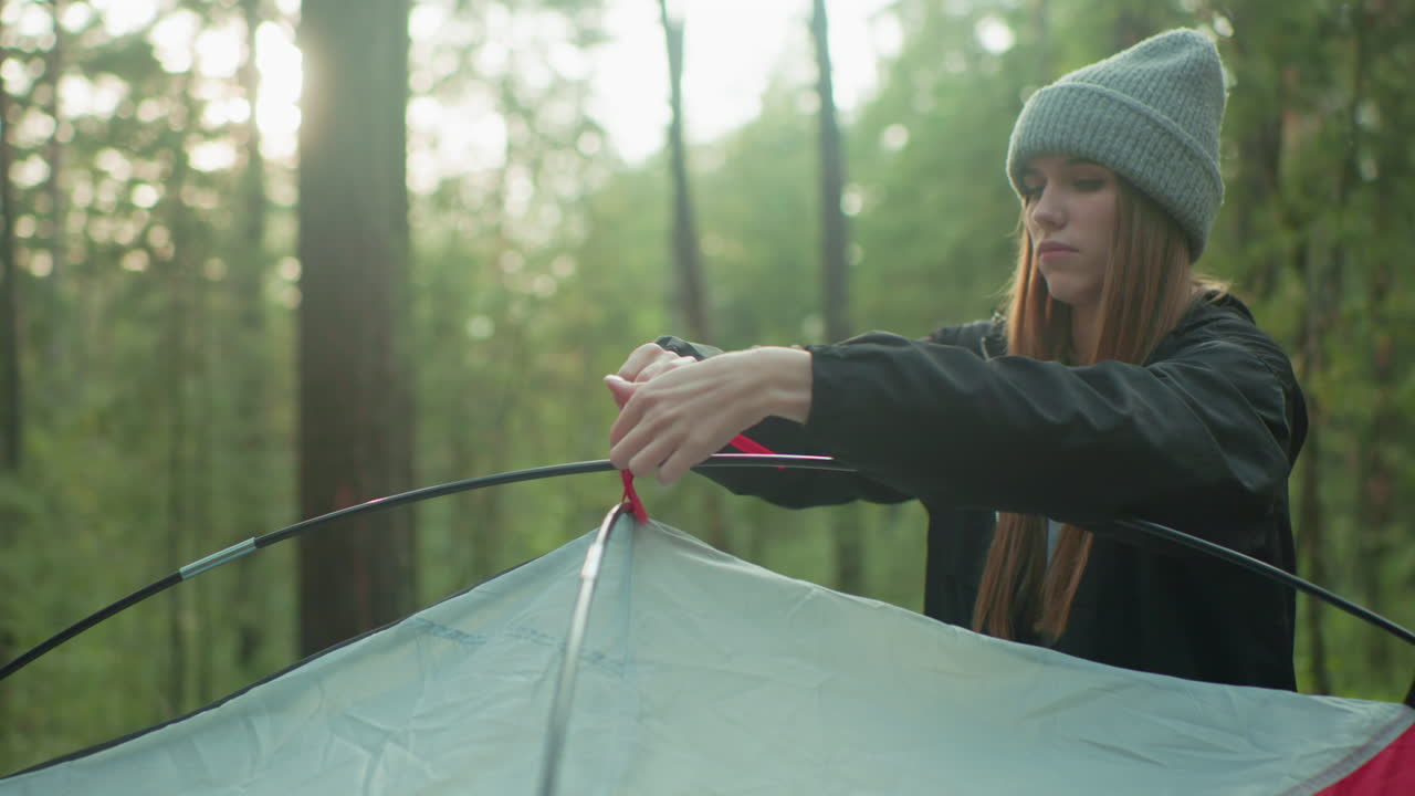 student wearing beanie calmly ties red rope to flexible tent pole while setting up shelter in forest environment during early morning outdoor activity surrounded by green trees and soft light