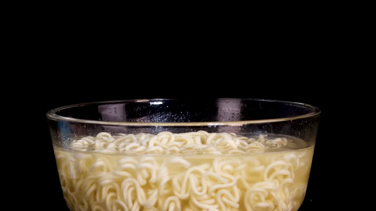 A hand sprinkles spice powder onto a bowl of instant noodles in clear broth, shot against a black background with dramatic studio lighting and static camera