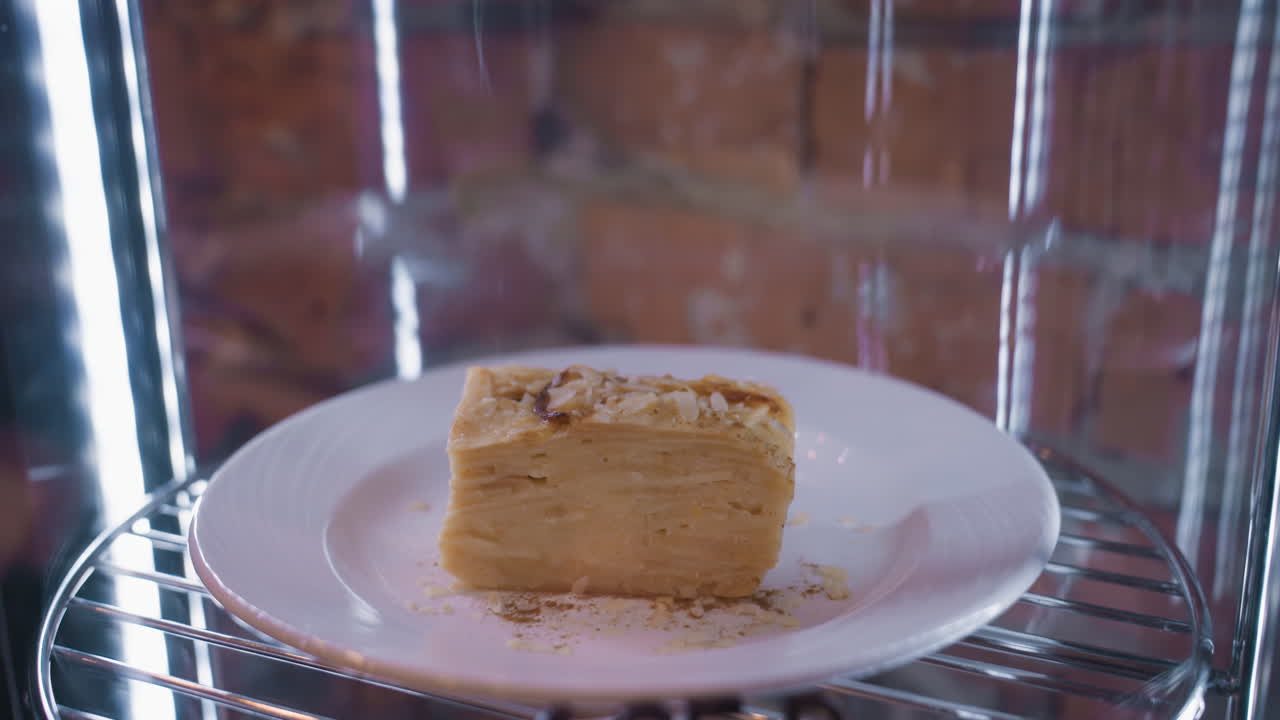 Close-up view of dessert roll slice on white plate placed inside glass display with visible light reflections and metal rack. Sweet treat appears layered, garnished with flakes and powdered topping