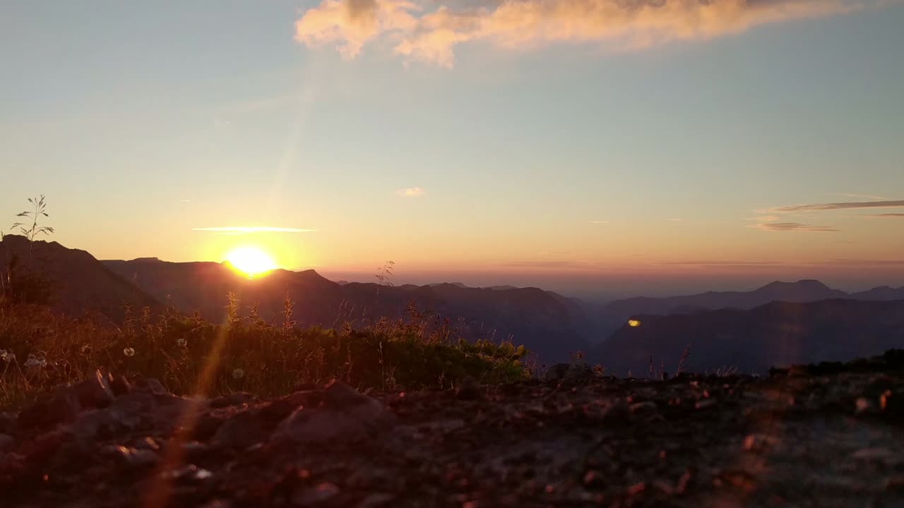 puesta de sol en la región alpina, el sol naranja se pone en el horizonte, la hierba y las flores soplan en el viento, la cámara está en un ángel bajo cerca del suelo, las nubes en el cielo