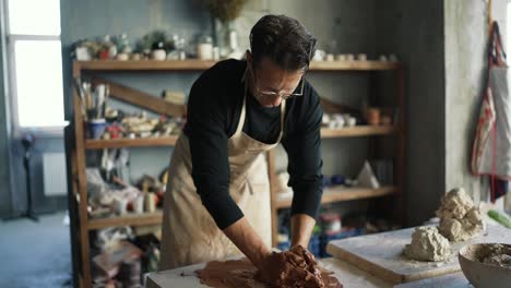 a happy young potter in glasses and and apron is crumpling a piece of clay on a special board in his studio. traditional art