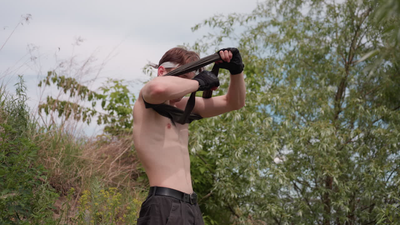 Warrior in cargo pants attempts to put on sleeveless shirt while murmuring words alone outdoors near trees and slope under cloudy sky, with tactical gloves on hands and determined expression on face