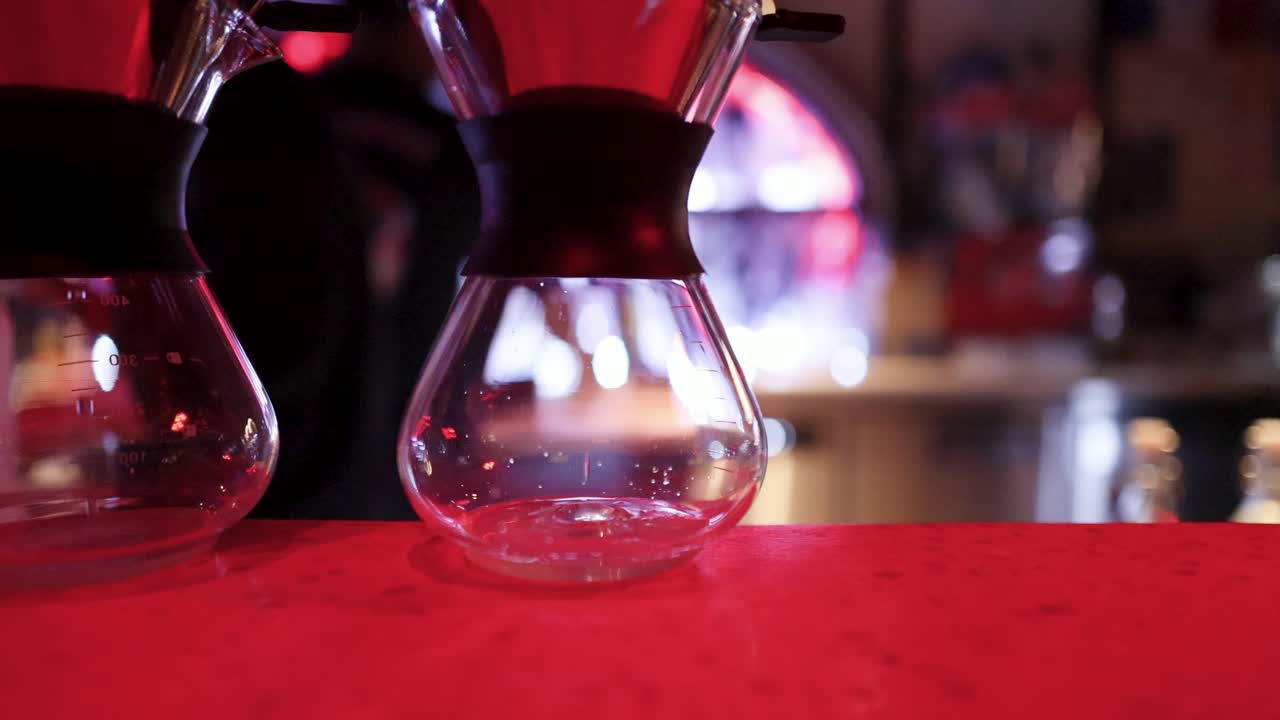 Barista pours liquid into glass Chemex coffee maker under moody, colorful lighting in Bangkok bar