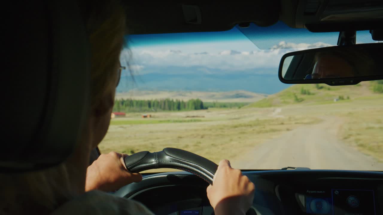 Pessoa dirige veículo por estrada de terra rural através de campos abertos. Céu azul claro com nuvens esparsas. Motorista focado, mãos firmes no volante. A paisagem se estende ao longe com gramíneas e árvores distantes.