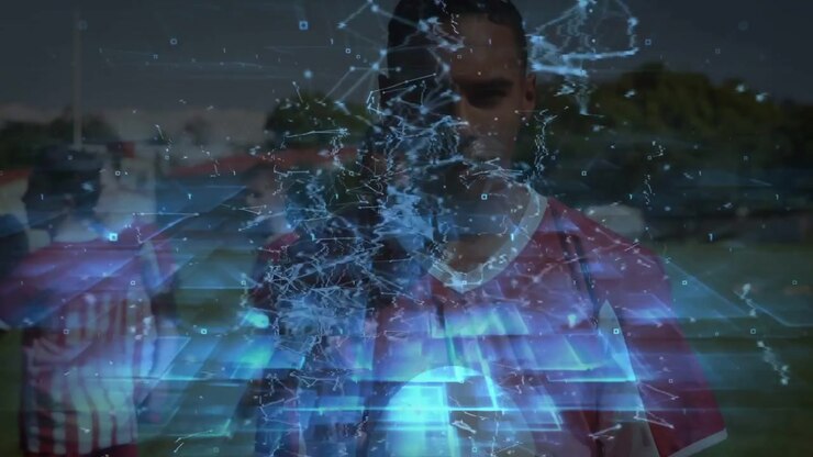 Homem adulto segurando uma bola de futebol, time em pé no campo, com uma sobreposição holográfica azul surgindo mostrando estatísticas de futebol.