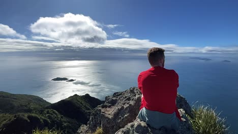 Redhead-male-hiker-looks-out-at-ocean-with-fluffy-white-clouds,-New-Zealand