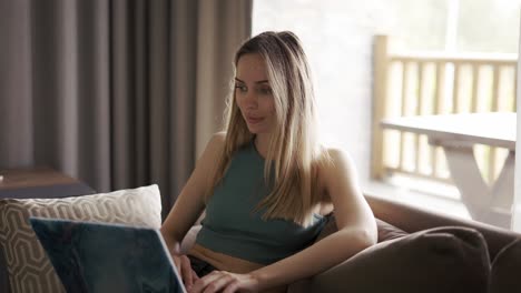 Blonde-woman-sitting-on-sofa-in-country-house,-browsing-social-media-on-laptop