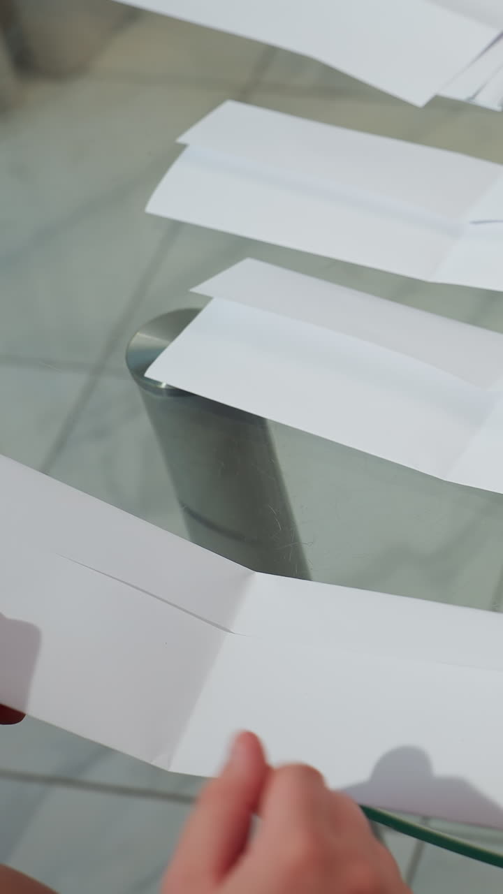 Niño repitiendo dobleces de papel sobre una mesa de cristal; primer plano de unas pequeñas manos colocando varias tiras y doblando filas pulcras; práctica paciente de una secuencia básica tipo origami para desarrollar la habilidad de doblado.