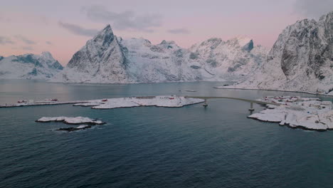 Moskenesøya-snowy-mountain-skyline-low-aerial-view-establishing-Reine-Norway-fishing-village-at-sunrise