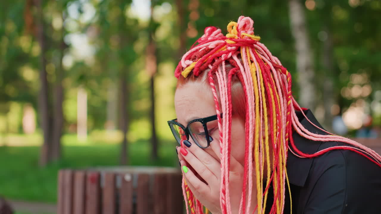 Mujer blanca sentada en un banco, inclinada hacia delante, con las manos sobre la boca, rastas llamativas, papelera en un parque urbano cercano, luz del atardecer, expresión tensa y de malestar que sugiere náuseas o shock