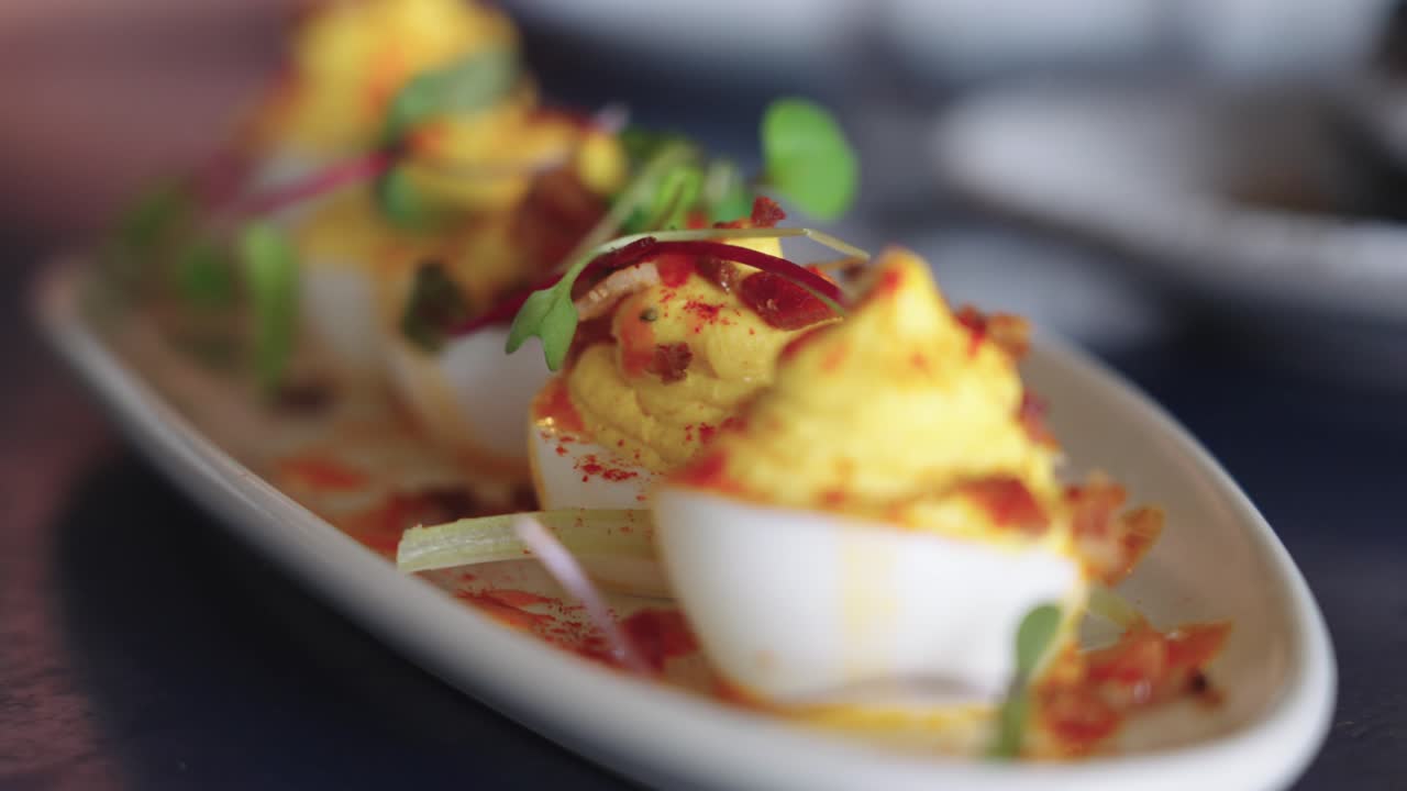 Filmato 4K al rallentatore di delicate uova ripiene guarnite con micro-verdure, servite su un piatto ovale bianco in un elegante ristorante per brunch, evidenziando una presentazione gourmet.