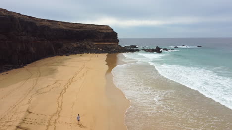 Wave-washed-sandy-beach-Escalera-with-a-few-visitors-relaxing-on-the-sea-shore-with-dark-rocky-cliffs-on-an-overcast-day,-forward-aerial-shot