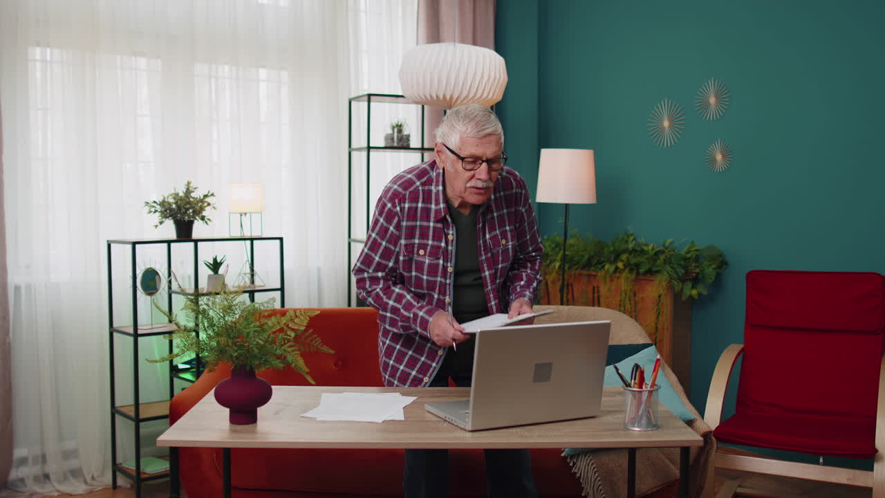 Senior grandfather man working on laptop computer making notes on sheet of paper calculating bills