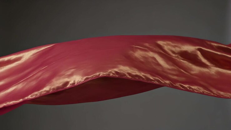 Tessuto di raso rosso ondeggiante che si muove attraverso uno studio grigio, con un ventilatore fuori campo che soffia per uno studio sull'illuminazione.