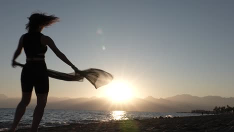 silueta de mujer joven en bikini con chal bailando y girando en la playa con puesta de sol sobre el mar