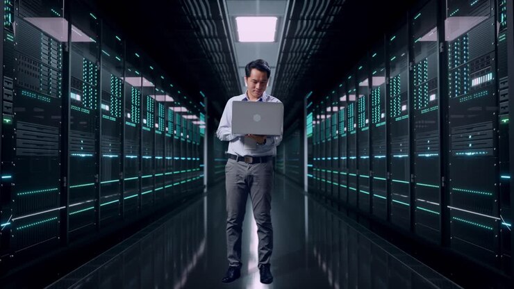 Helkropsbillede af en asiatisk mandlig professionel medarbejder, der står med sin bærbare computer i et serverrum i et datacenter og arbejder uafbrudt.