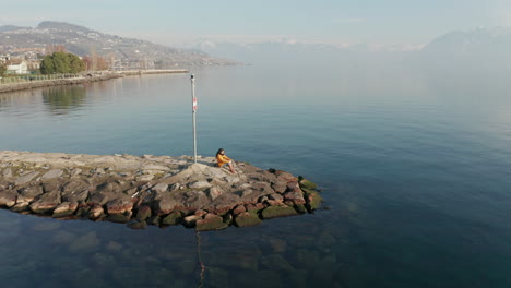 Aerial-of-woman-sitting-at-the-end-of-quay-at-beautiful-lake-with-mountains-in-the-far-background