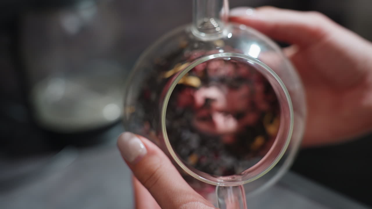 CloseUp Hands Placing Dried Herbs Into Glass Teapot, Artisan Herbalist Preparing Loose Leaf Infusion On Countertop With Petals And Measuring Focus, Transparent Vessel Highlights Texture And Color