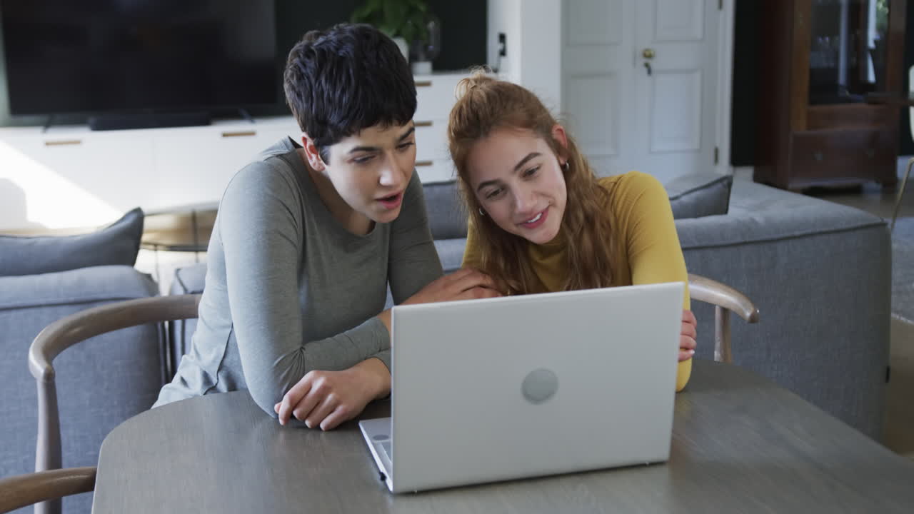 una feliz pareja de lesbianas caucásicas sentadas, sonriendo y usando una computadora portátil en la sala de estar soleada