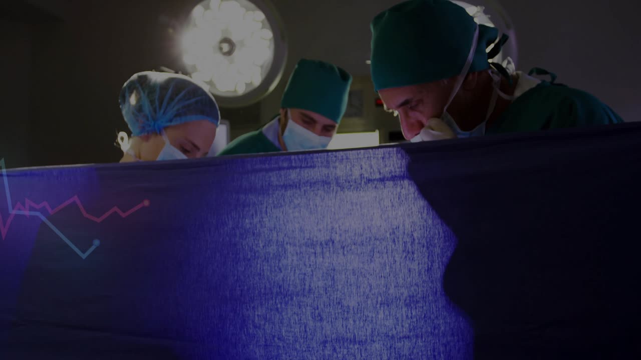 Blå duk läggs under operationslampa, team justerar verktyg medan de övervakar en vitalparametergraf.