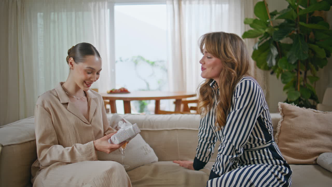 Two women exchanging gifts sitting on couch living room with happy smiles