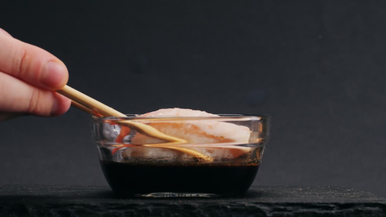 Hand holding chopsticks, picking up shrimp sushi from a bowl of soy sauce