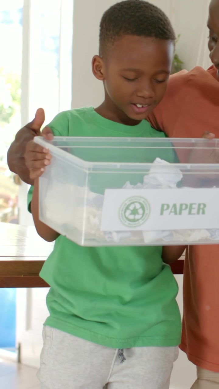 Vertical video: Holding PAPER bin, green-tee boy looking at adult in orange after prompt at home