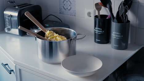 medium shot of a steaming pasta tagliatelle in a big steel pot with two wooden spoons and a plate on the side in a white kitchen surrounded by kitchen utensils during a day