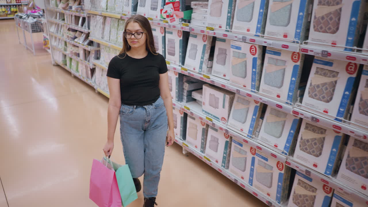 vista trasera de una mujer joven con camiseta negra y vaqueros caminando por el pasillo de la tienda, navegando por productos de cama mientras sostiene bolsas de compras, otro comprador pasa en el fondo