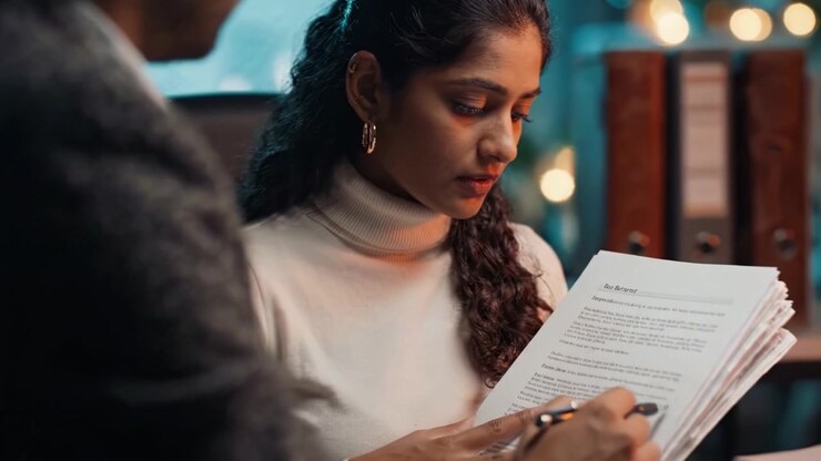 A focused woman reviews important documents with a thoughtful expression in a well-lit office, highlighting the significance of hard work and detail in her task.