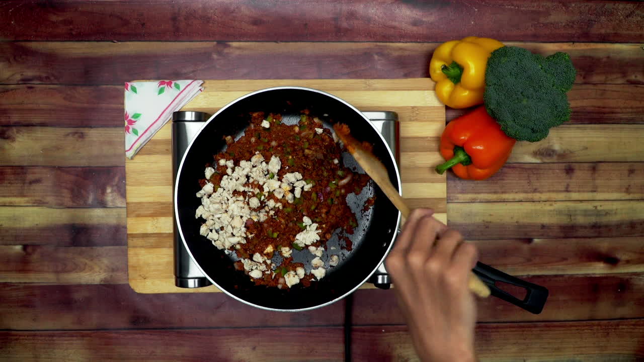 una vista superior de la adición de pollo a los ingredientes en la sartén, colocados en una estufa, dos grandes pimientos amarillos y rojos y un brócoli verde sobre la mesa