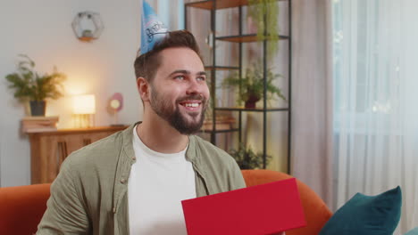 un joven feliz y sorprendido usa un sombrero de cumpleaños festivo abriendo una caja de regalos en el sofá del sofá en casa