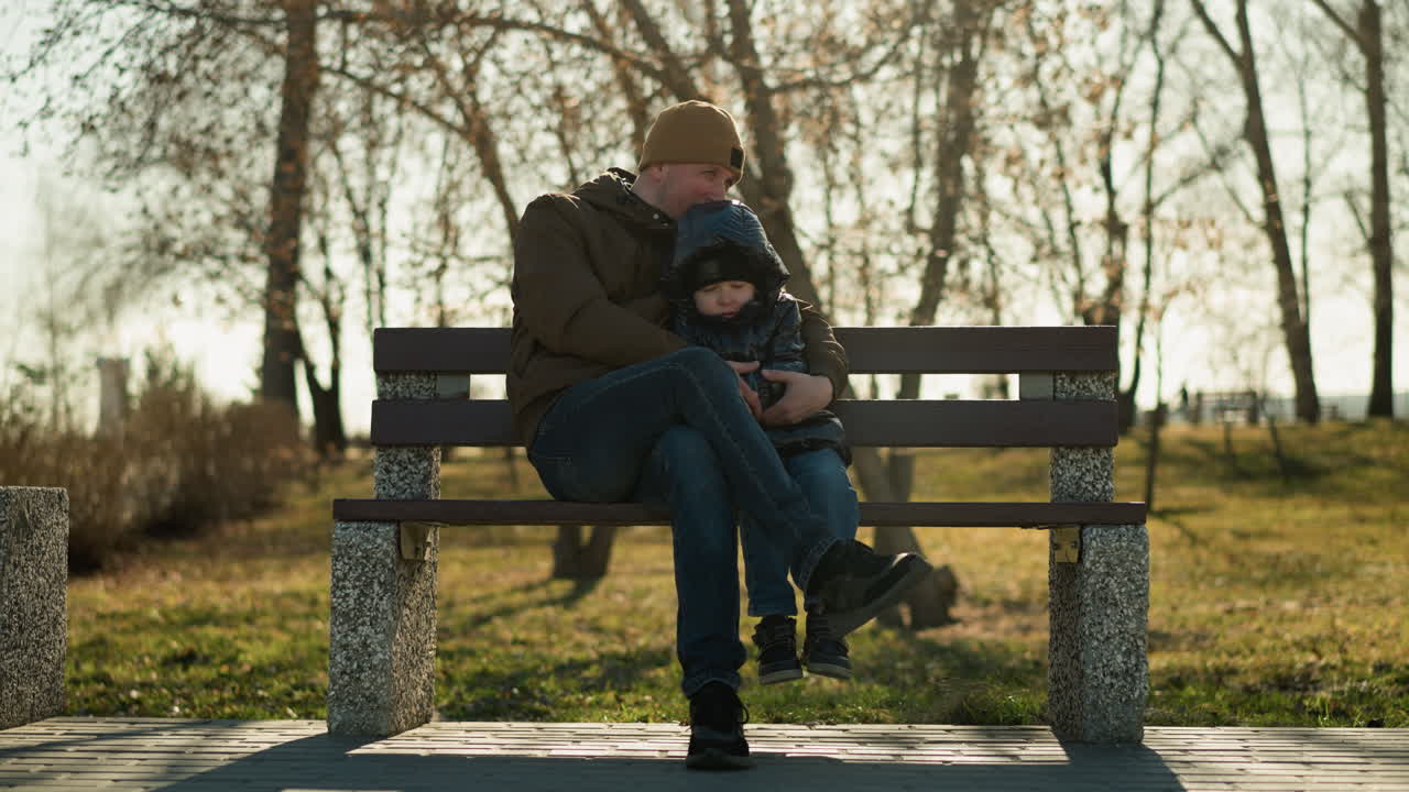 Father and son sitting on a bench in a park, the father rests his head gently on his son's head as he holds him close with leg crossed, with tress in the background