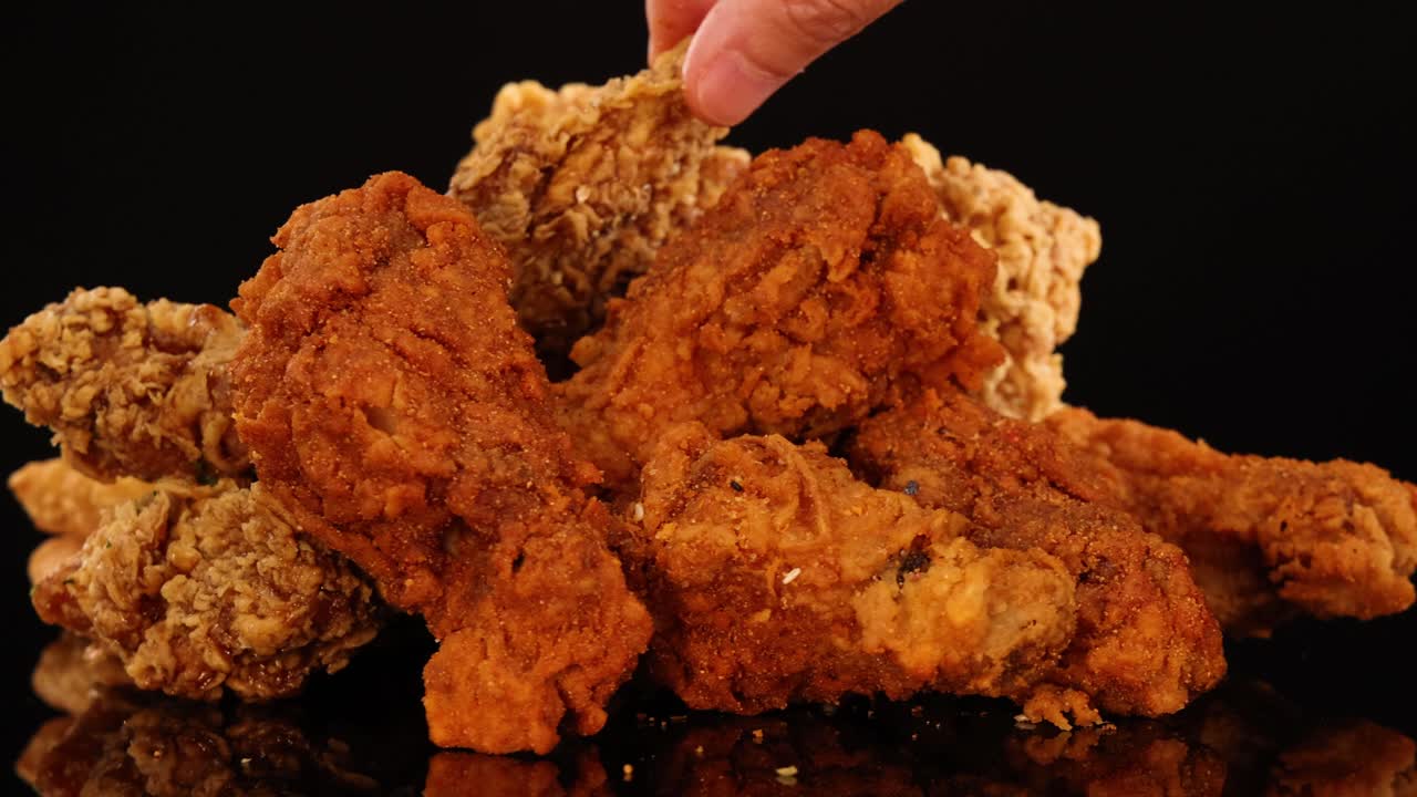 A hand reaches into a pile of assorted crispy fried chicken wings, drumsticks, and nuggets under studio lighting with a black background, highlighting texture and color