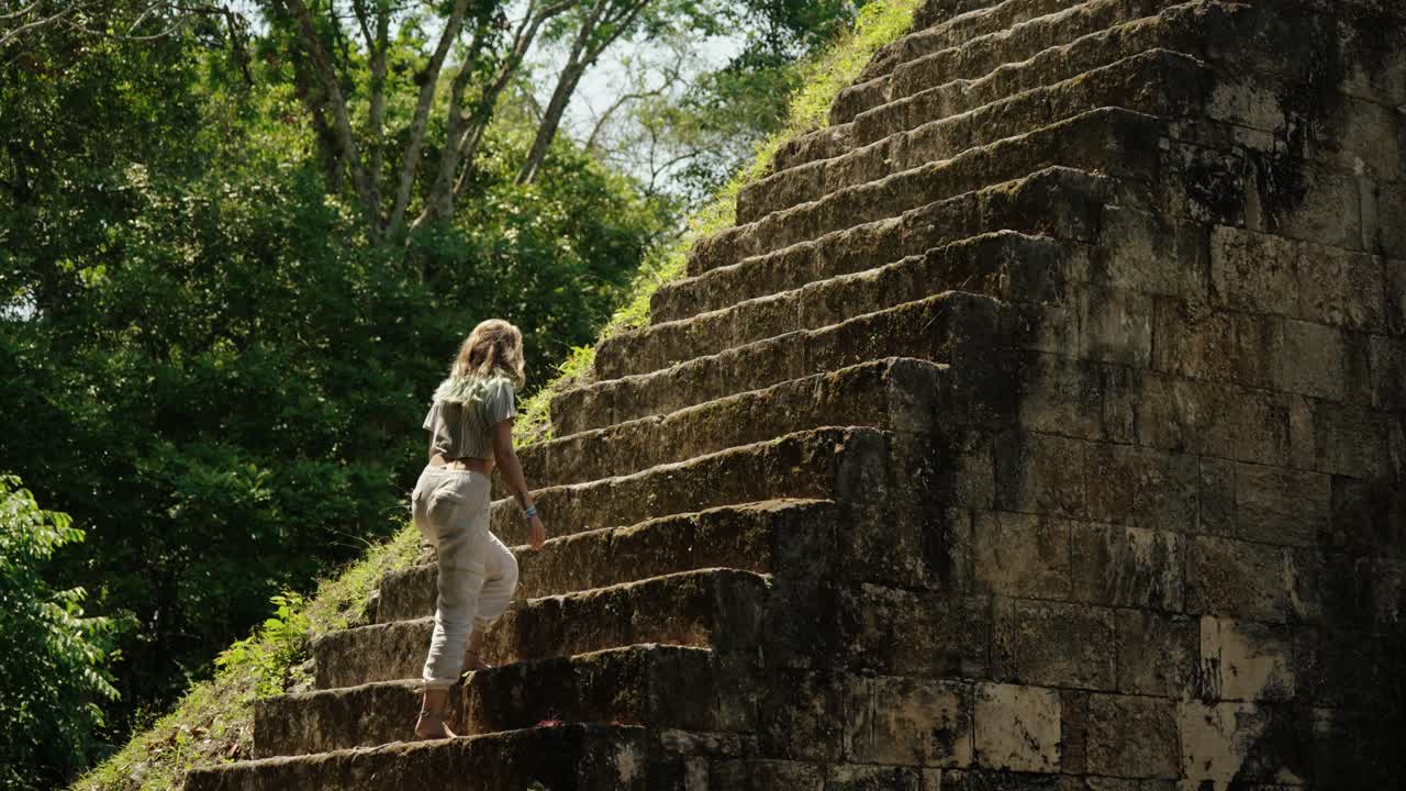 Woman climbs an ancient Mayan pyramid in Tikal, Guatemala, surrounded by jungle and sunlight filtering through the trees.