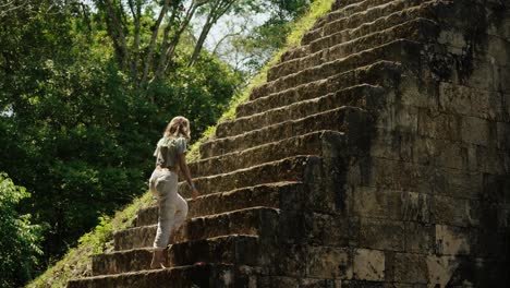 woman climbs an ancient mayan pyramid in tikal, guatemala, surrounded by jungle and sunlight filtering through the trees.