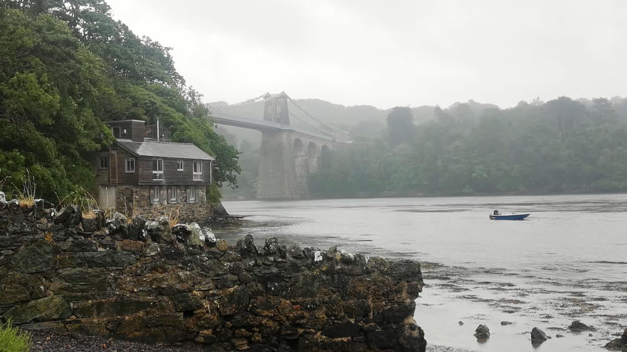 lluvia brumosa a través de la pintoresca casa de vacaciones en la orilla del río bajo el puente del estrecho de menai, anglesey