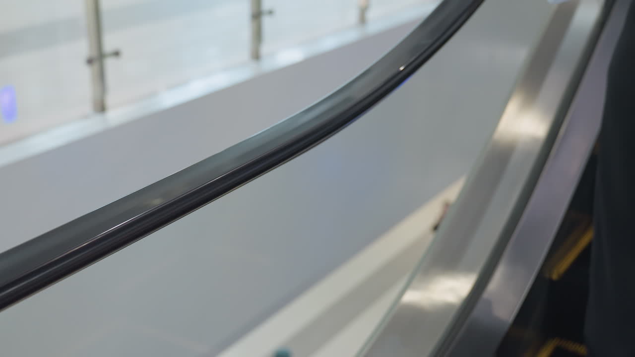Close up side view of girl riding moving escalator with focus on handrail and metal edge, while blurred background reveals mall interior and indistinct people below