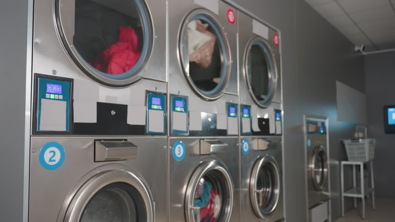 Side view labelled steel washing machines in row spinning clothes, digital timers glowing, modern laundromat interior, numbered doors, stainless panels, clean facility, active wash cycle behind glass