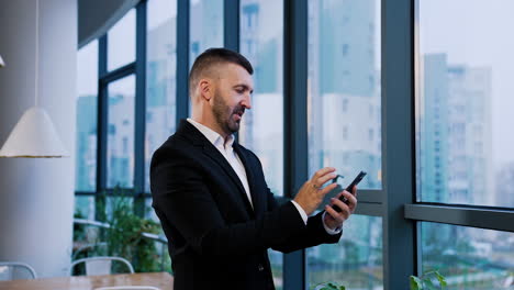dark-haired bearded man wearing suit stands at window. businessman looking for information in his phone. cityscape at backdrop in blur.