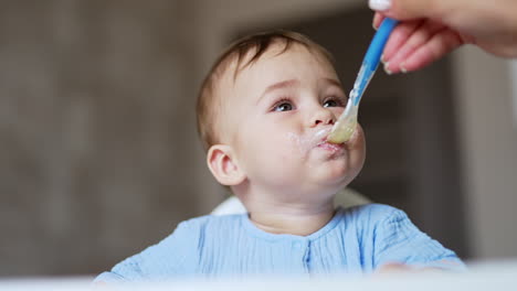 happy baby boy is fed from spoon by his mom. lovely kid eating and tasting food. close up. blurred backdrop.