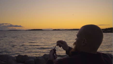fumador profesional sentado junto al mar en una playa rocosa arreglando su pipa con una herramienta para tener una mejor experiencia de fumar solo sin amigos independiente sigma masculino maravilloso cielo amarillo naranja